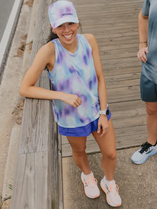 A girl standing against a wooden railing outside wearing a tie dye hat and matching tie day shirt with purple shorts and orange shoes